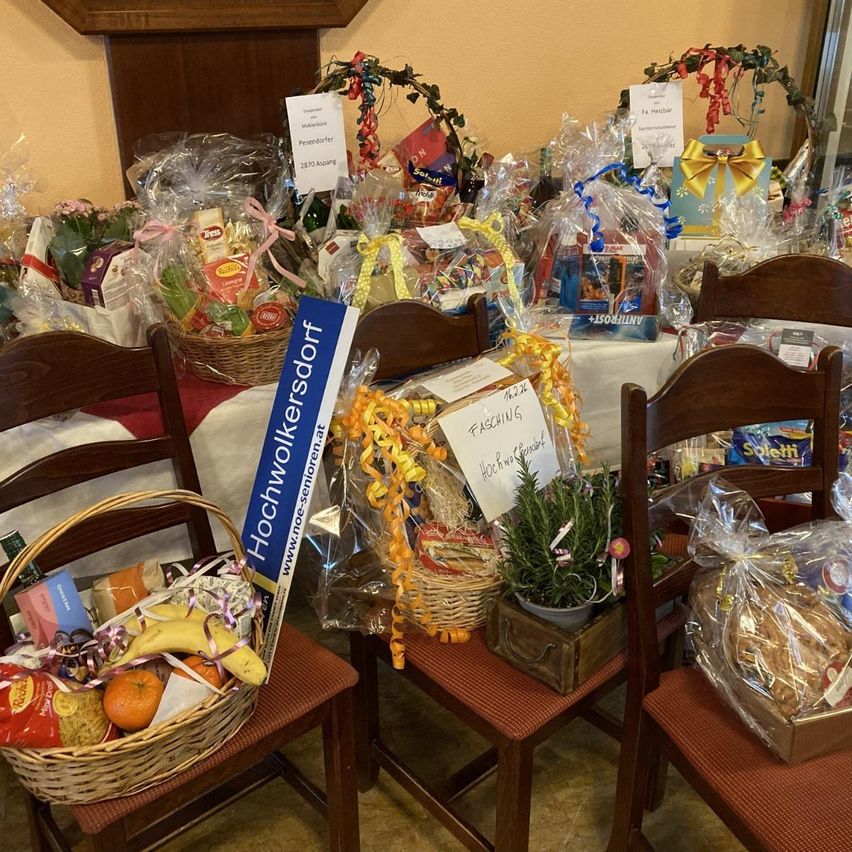 Verschiedene Geschenkkörbe, einige in Plastik eingewickelt, sind auf Stühlen um einen Tisch herum angeordnet. Jeder Korb enthält verschiedene Gegenstände wie Obst, Snacks und einen Topf mit Pflanzen. Ein Schild lautet 'Hochwolkersdort'.