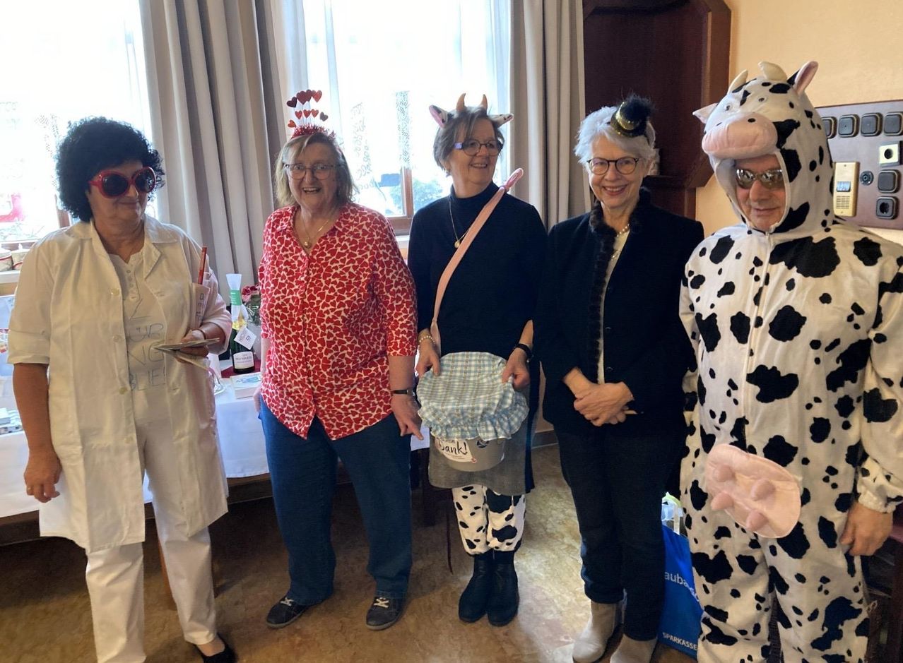 Fünf Frauen stehen in einem Raum, alle in Kostümen gekleidet. Zwei tragen Kuhkostüme, eine ein Hexenkostüm und zwei andere Kostüme. Sie lächeln für ein Foto.
