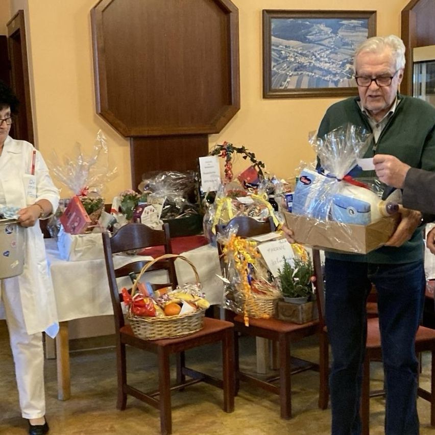 Ein älterer Mann hält einen Geschenkkorb, während eine Frau in einer weißen Jacke einen Eimer hält. Ein Tisch mit verschiedenen Geschenkkörben und einer Pflanze ist im Hintergrund.