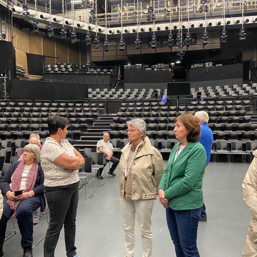 Eine Gruppe von Frauen steht in einem leeren Theater, mit Reihen von Stühlen und Bühnenbeleuchtung sichtbar.
