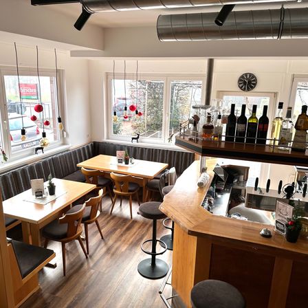 Ein Innenrestaurant mit Holzboden, einer Bartheke und einem großen Fenster mit einer Hirschsilhouette. Die Bartheken hat Flaschen mit Schnaps und eine Uhr.