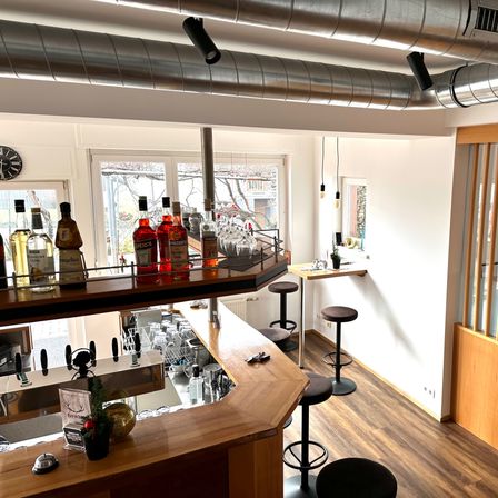 Innenansicht einer Bar mit einer Holztheke, Regalen mit Flaschen und schwarzen Hockern. Ein Fenster und Weihnachtsdekoration sind ebenfalls zu sehen.