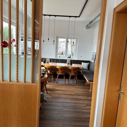 Ein Essbereich mit Holzstühlen, einem langen Tisch und einer Bank mit einem Kissen. Der Raum hat weiße Wände, eine Glasabtrennung und hängende Lampen. Durch das Fenster draußen sind Bäume und eine Aussicht auf das Äußere zu sehen.