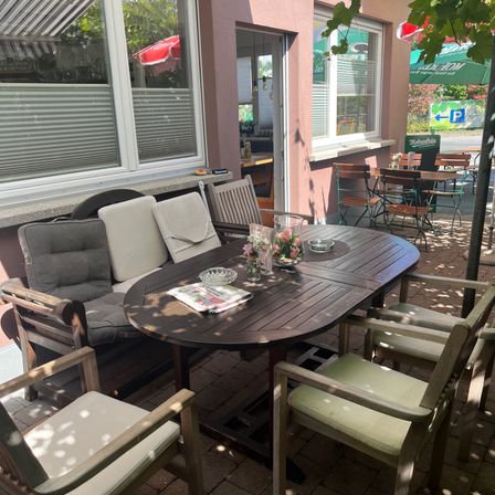 Ein Holztisch mit Blumen und einer Zeitung steht in einer Außensitzecke mit einer Couch und Stühlen. Der Bereich ist von einem Ziegelboden und einem Gebäude mit Fenstern und einer Tür umgeben.