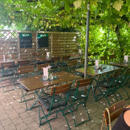 Ein Gartenrestaurant mit Holztischen und Stühlen, umgeben von Grün und einer Holzzaun. Es gibt Tafeln mit Schrift auf dem Zaun.