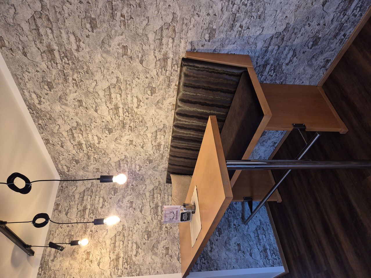 Ein Nahaufnahme einer modernen Inneneinrichtung mit einer strukturierten Wand, einer Holzbank und einem Tisch mit einem Menü. Zwei Lampen hängen von der Decke.