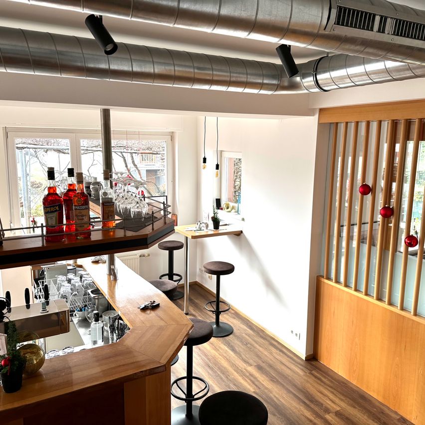 Ein Barbereich mit Holzboden und Theke. Regale mit Flaschen und Gläsern sowie rote Ornamente, die an einer Holztrennwand hängen.