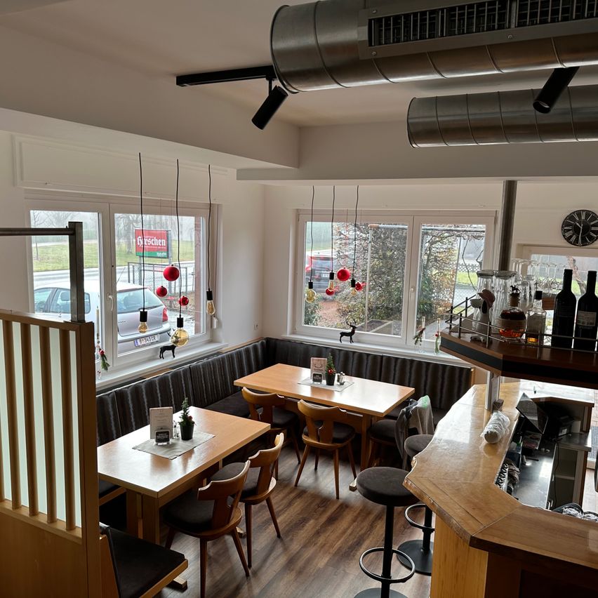 Ein Restaurantinnenraum mit Holztischen, Stühlen und einer Bartheke. Der Raum hat große Fenster mit Blick auf ein Auto und Bäume draußen. Es gibt Lampen, eine Uhr und einige Dekorationen.