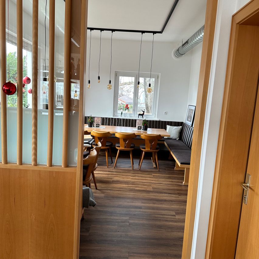 Ein Innenraumblick auf einen Essbereich mit Holzboden, einem langen Tisch, Stühlen und einer Bank. Es gibt hängende Lampen und Weihnachtsdekorationen an der Wand.