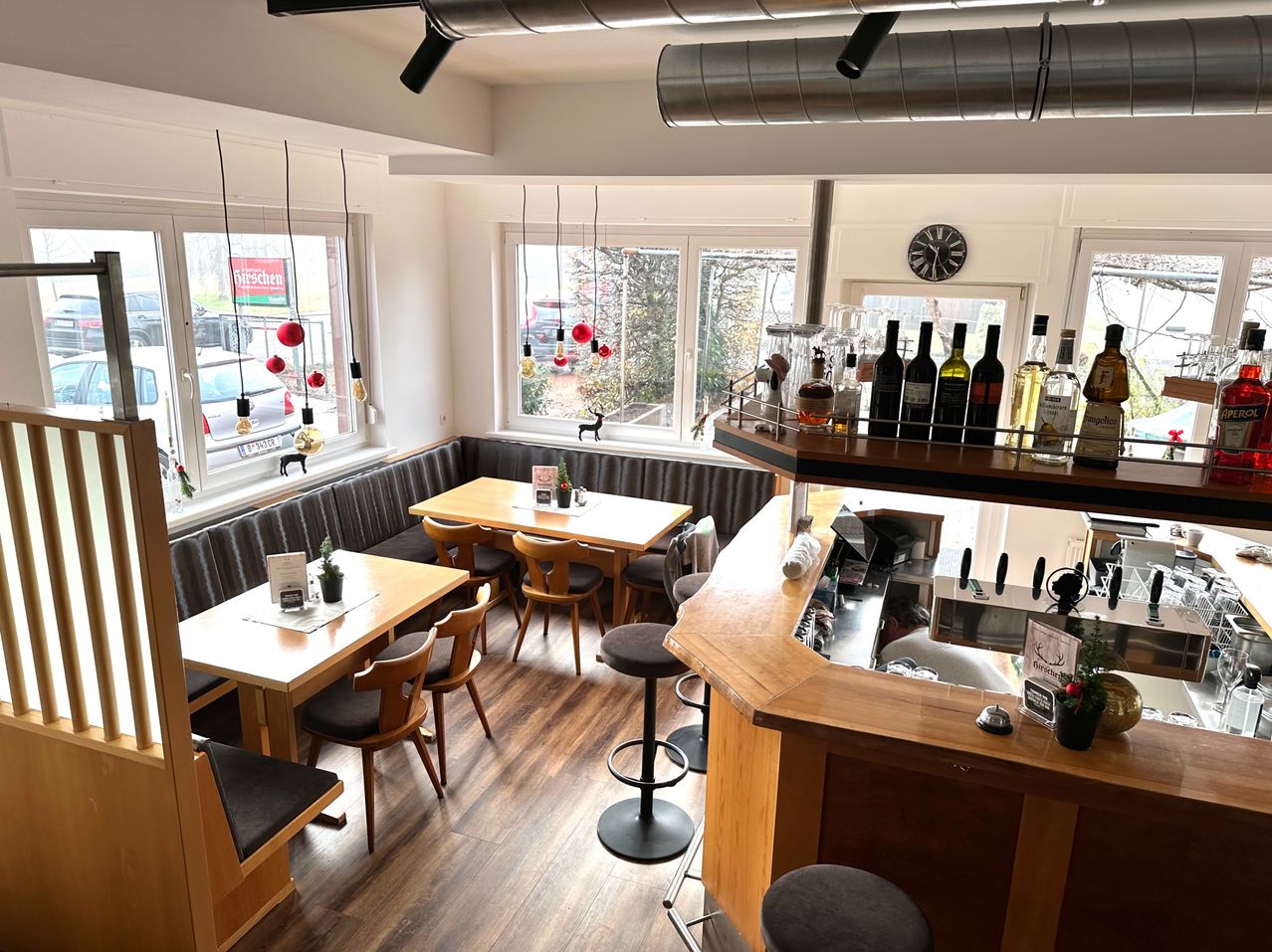 Ein Restaurant mit Holzböden und Tischen. Eine Bar mit einer Theke und Hockern. Flaschen Wein sind auf einem Regal ausgestellt. Eine Uhr ist an der Wand montiert.