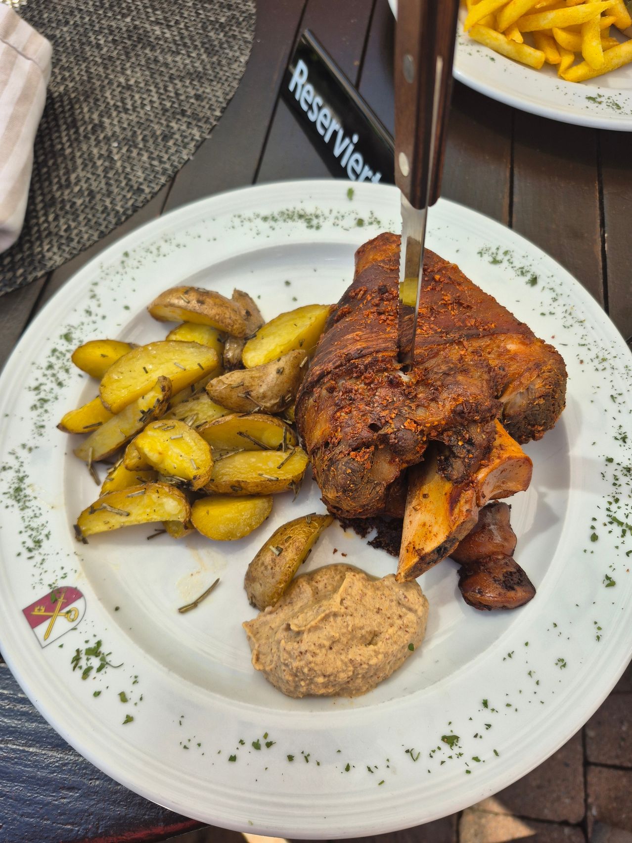 A white plate with roasted leg of lamb, potatoes, and a dip. The meat has a crispy crust and the potatoes are golden brown.