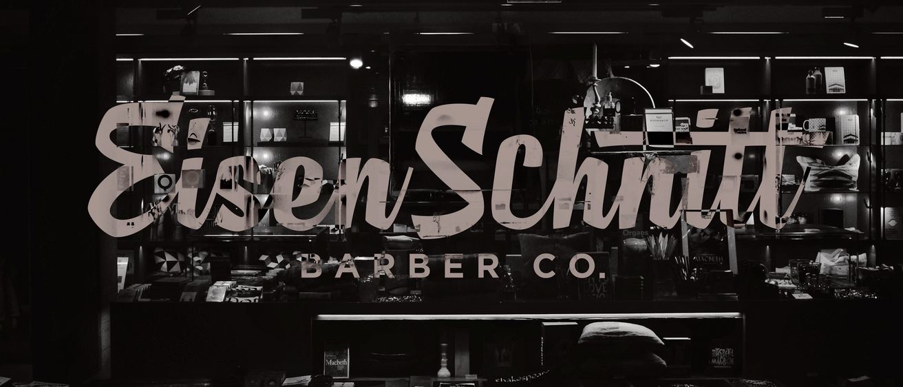 Ein Schwarz-Weiß-Bild eines Barbierladenschilds mit dem Text 'Schen/Schin BARBER CO.' auf einem Fenster. Im Hintergrund sind Gegenstände auf den Regalen zu sehen.