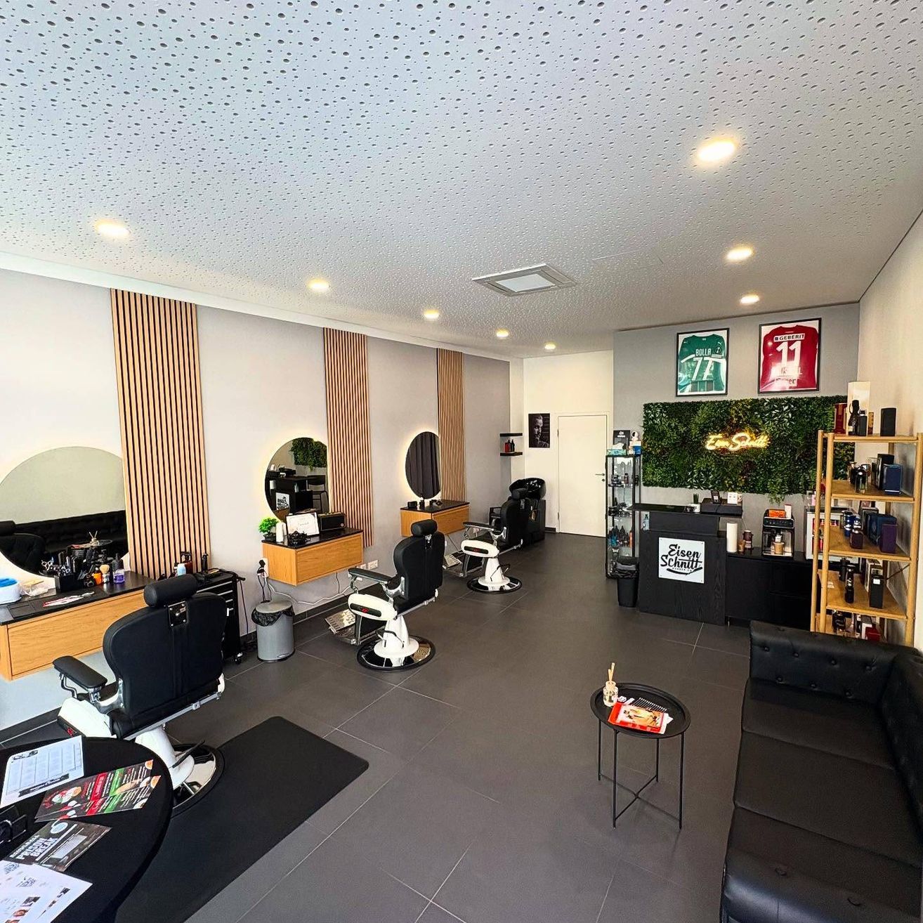 Ein moderner Friseursalon mit grau gefliesten Böden, schwarzen Ledersofas, weißen Wänden und Holzakzenten. Mehrere Friseurstuhle sind angeordnet, mit einem Mülleimer, einem Schreibtisch und einer Pflanze in der Nähe. Gerahmte Trikots hängen an der Wand.