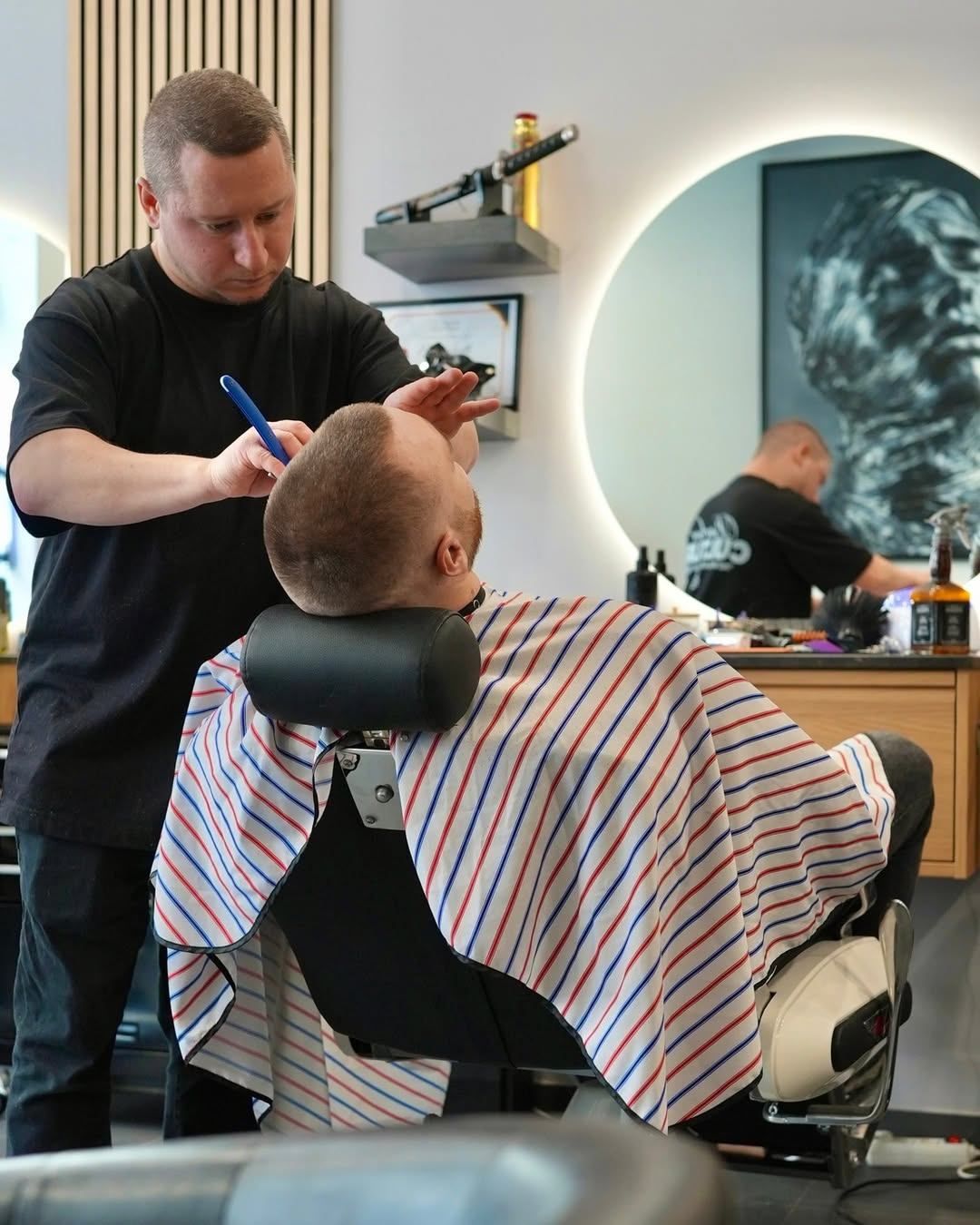 A barber in a black shirt trims a man's hair with a blue razor, with a mirror and a picture in the background.