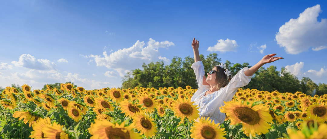 Eine Frau steht freudig in einem Sonnenblumenfeld unter einem klaren blauen Himmel mit ausgestreckten Armen. Sie trägt ein weißes Oberteil und eine Blumengirlande im Haar.