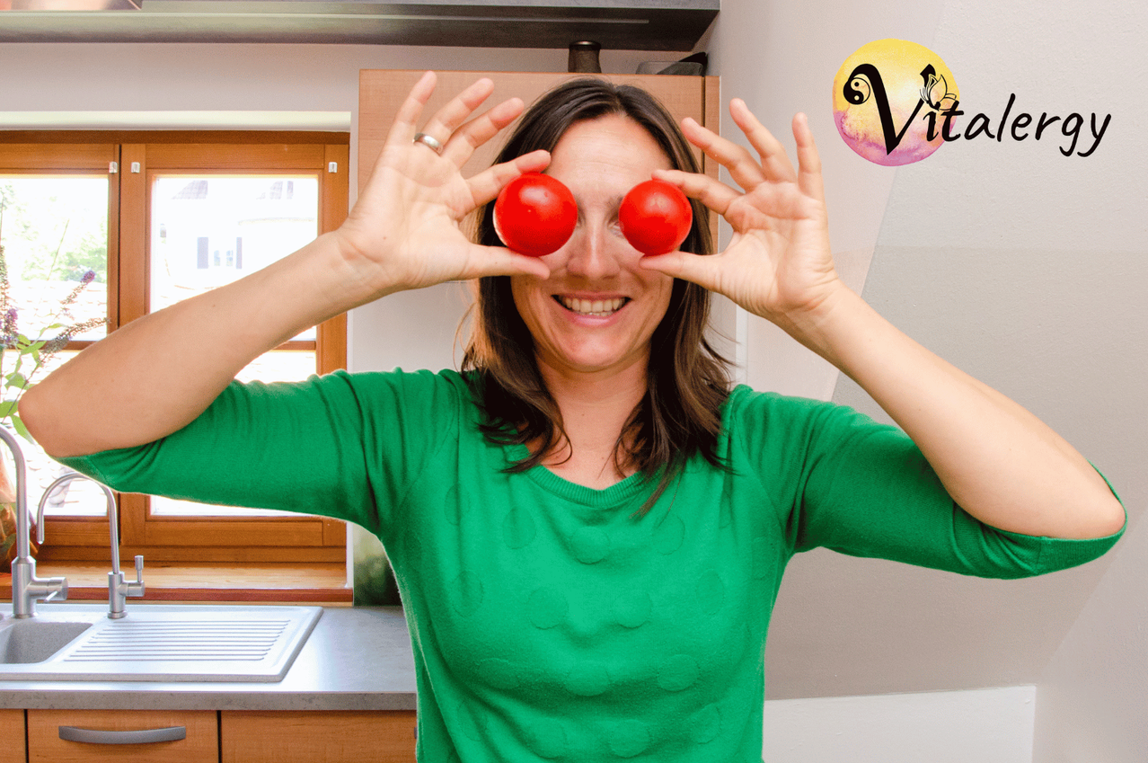 Eine Frau in einem grünen Shirt lächelt und hält zwei rote Tomaten über ihren Augen. Hinter ihr ist eine Küche mit Holzschränken und einem Fenster.