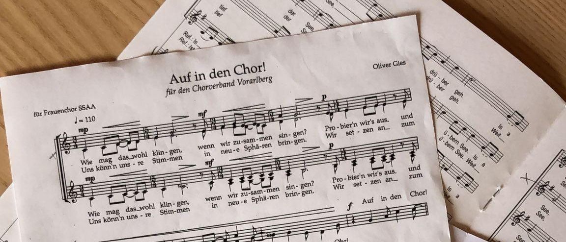 Partituren für den Chor sind gestapelt. Die Noten sind in Deutsch geschrieben und enthalten eine Mischung aus Texten und musikalischen Notationen. Das Blatt zeigt eine Tonart und Taktart, wobei einige Noten hervorgehoben sind.