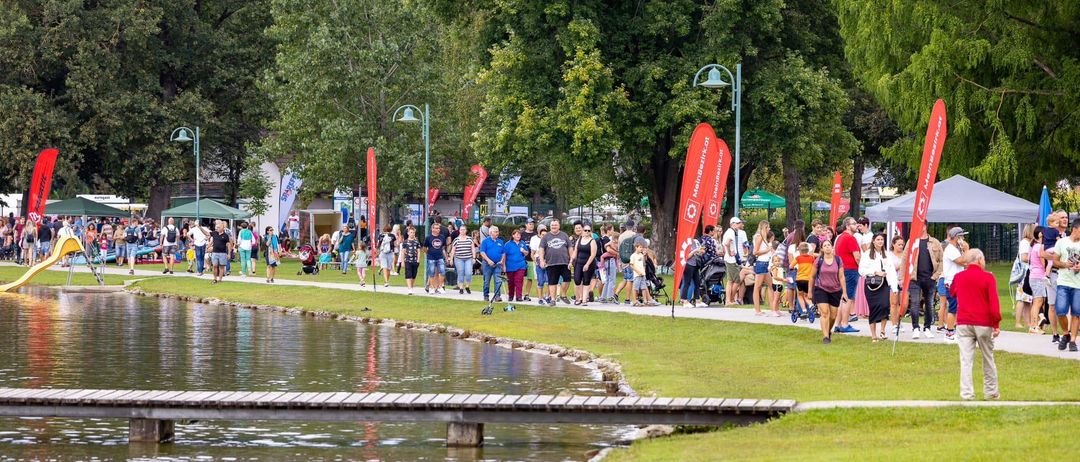 Eine große Menschenmenge, die entlang eines Seeufers mit Flaggen, Bäumen und einer hölzernen Brücke im Hintergrund spazieren geht.