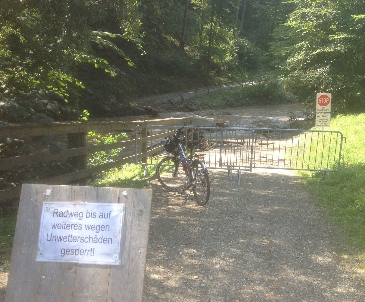 Ein Fahrrad steht am Rande einer Schotterstraße mit einem Stoppschild und einem geschlossenen Tor. Ein Schild steht, 'Radweg bis auf weiteres wegen Unwetterschäden gesperrt!' Bäume und Pflanzen umgeben das Gebiet.