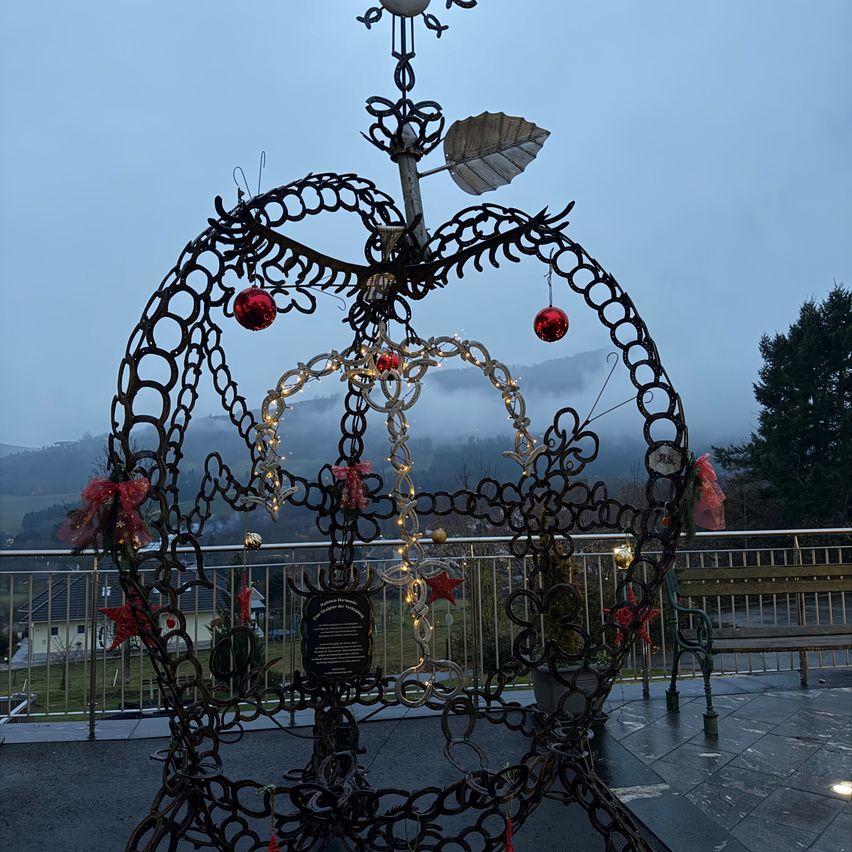 Eine komplizierte Schmiedeeisenkrone mit roten Ornamenten und Lichtern steht auf einem Balkon mit einem verschwommenen Bergblick im Hintergrund.
