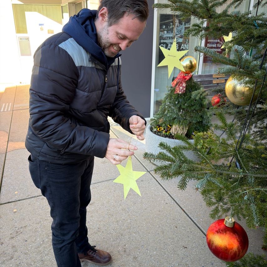 Ein Mann draußen in einer Jacke hängt einen gelben Stern an einen Weihnachtsbaum. Er lächelt und schaut nach unten. Ein kleinerer Baum steht neben dem Baum.