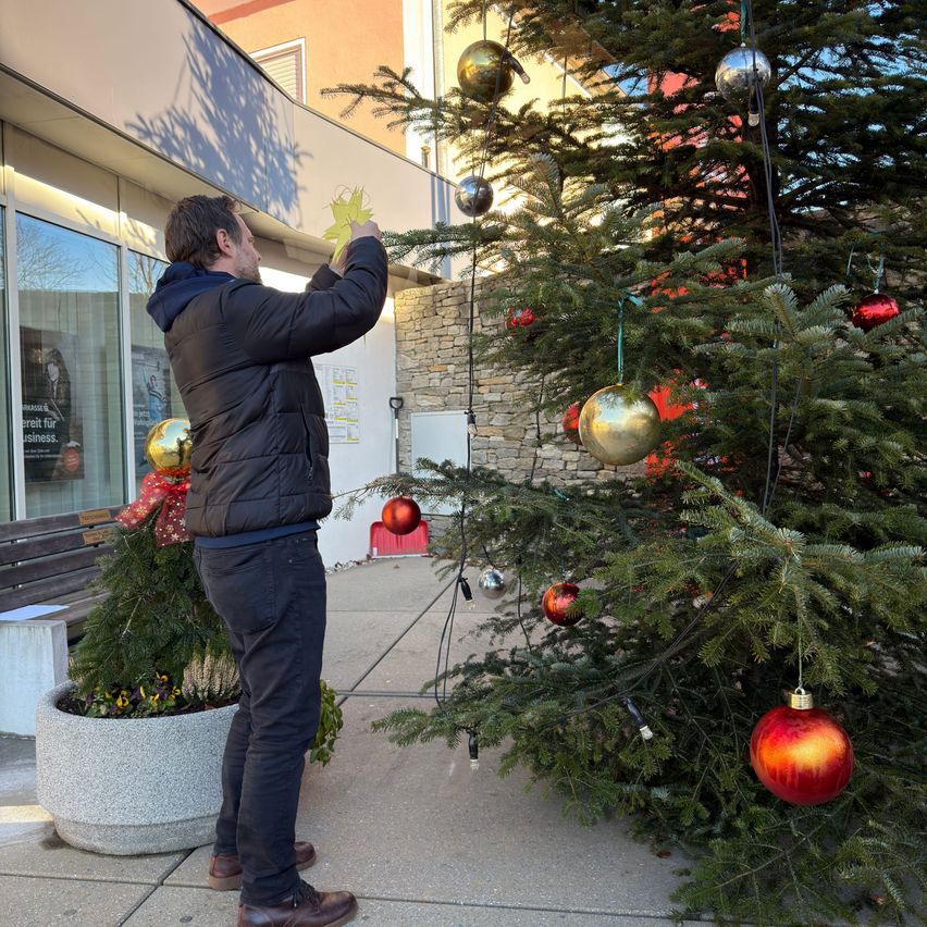 Ein Mann in einer Jacke schmückt eine Weihnachtsbaum vor einem Gebäude mit verschiedenen Ornamenten.