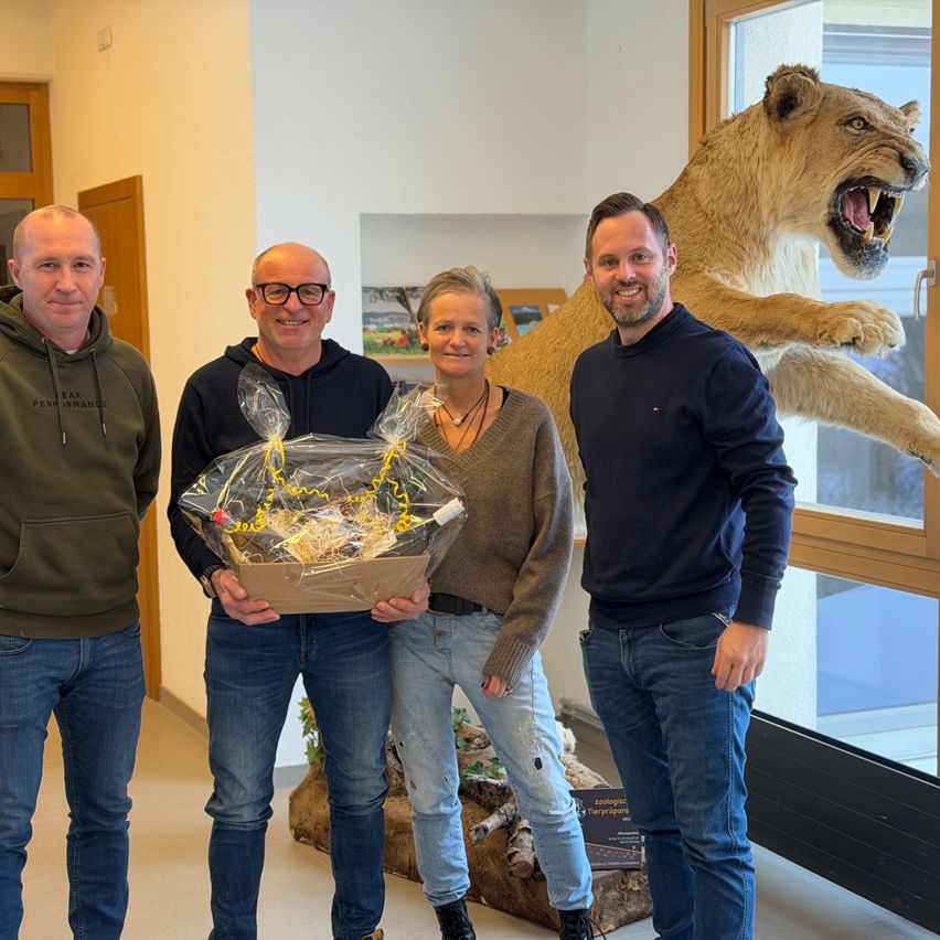Vier Personen stehen drinnen und lächeln. Der Mann links trägt eine grüne Kapuzenjacke. Der zweite Mann hält ein eingewickeltes Geschenk. Die Frau in der Mitte trägt eine Halskette. Der Mann rechts trägt ein dunkelblaues Hemd. Hinter ihnen steht eine große Löwenstatue.