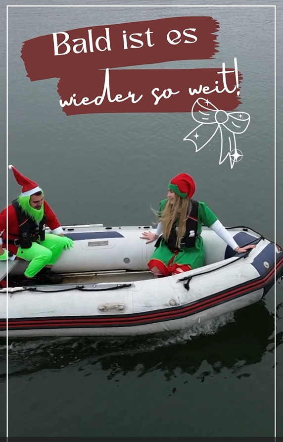 Zwei Menschen in Weihnachtskostümen auf einem Schlauchboot. Ein Mann trägt einen Weihnachtsmannhut und grüne Hose. Eine Frau trägt einen roten Hut und ein grünes Kleid. Das Boot hat rote und blaue Streifen.