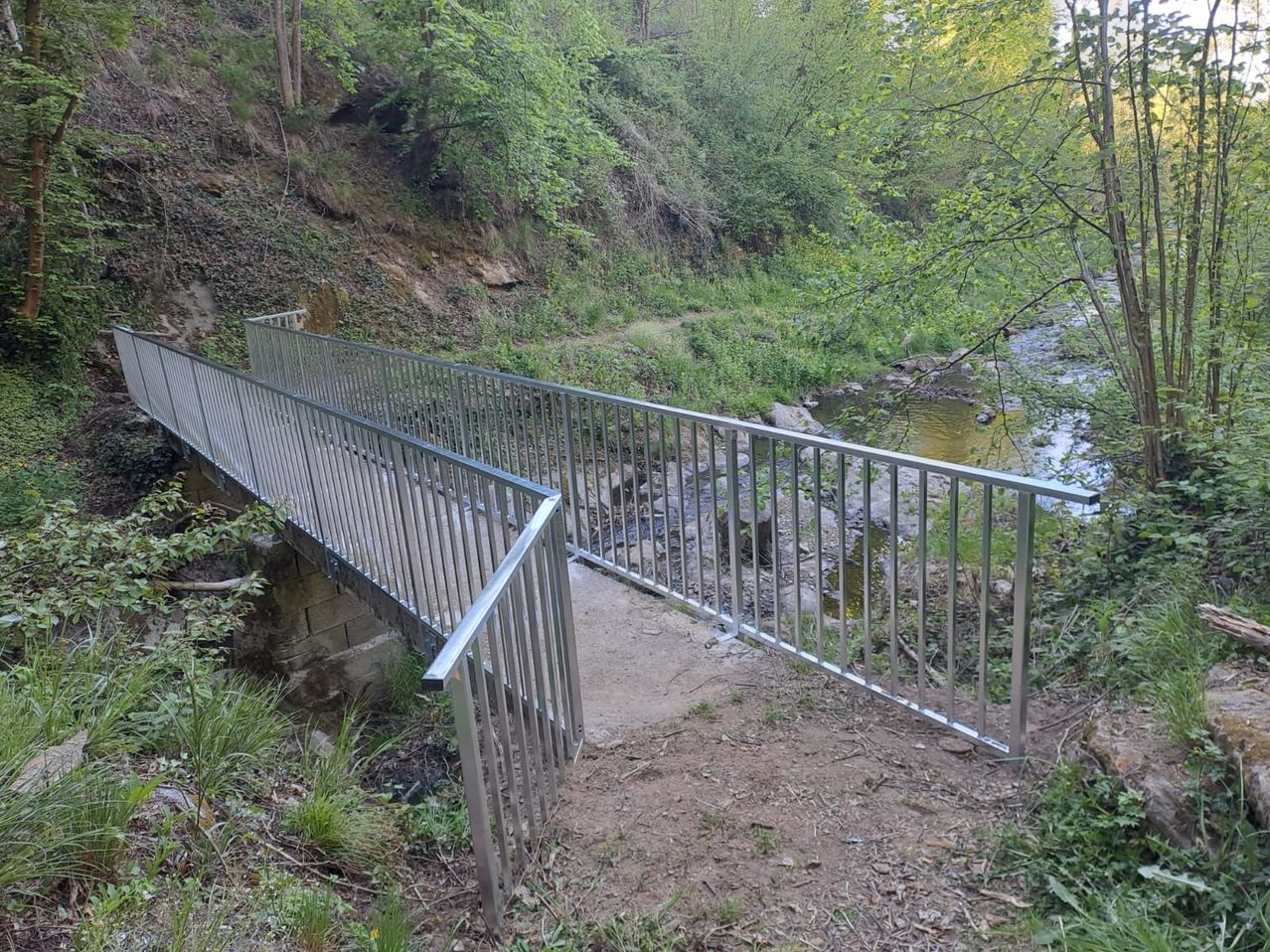 Eine Metallbrücke mit Geländern steht über einem Bach mit üppiger grüner Umgebung. Die Brücke ist robust und wirkt neu.