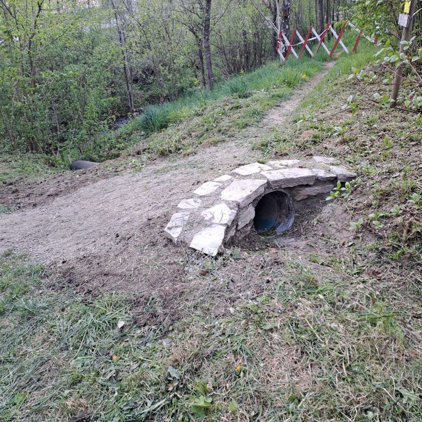 Ein mit Steinen bedeckter Durchlass mit einer Rohrleitung kommt an einem steilen Pfad aus dem Boden. Grüner Rasen und Bäume umgeben das Gebiet. Ein kleiner Bach fließt in der Nähe.