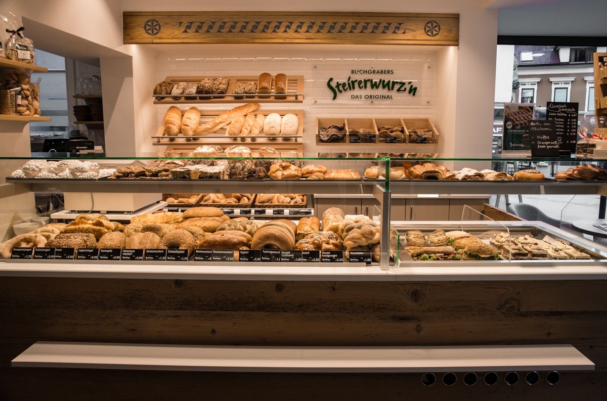 Eine Bäckerei-Vitrine mit verschiedenen Brot- und Gebäcksorten, einschließlich eines Schilds mit der Aufschrift 'Steirerwurzen Das Original'. Die Vitrine steht auf einer Holztheke.
