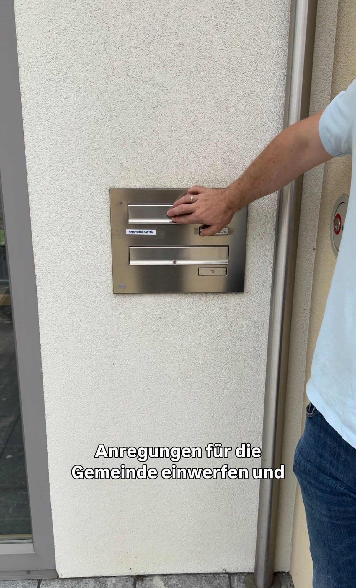 Ein Mann steckt einen Umschlag in einen silbernen Briefschlitz an einer weißen Wand, wobei die Worte 'Anregungen für die Gemeinde einwerfen und' oben sichtbar sind.