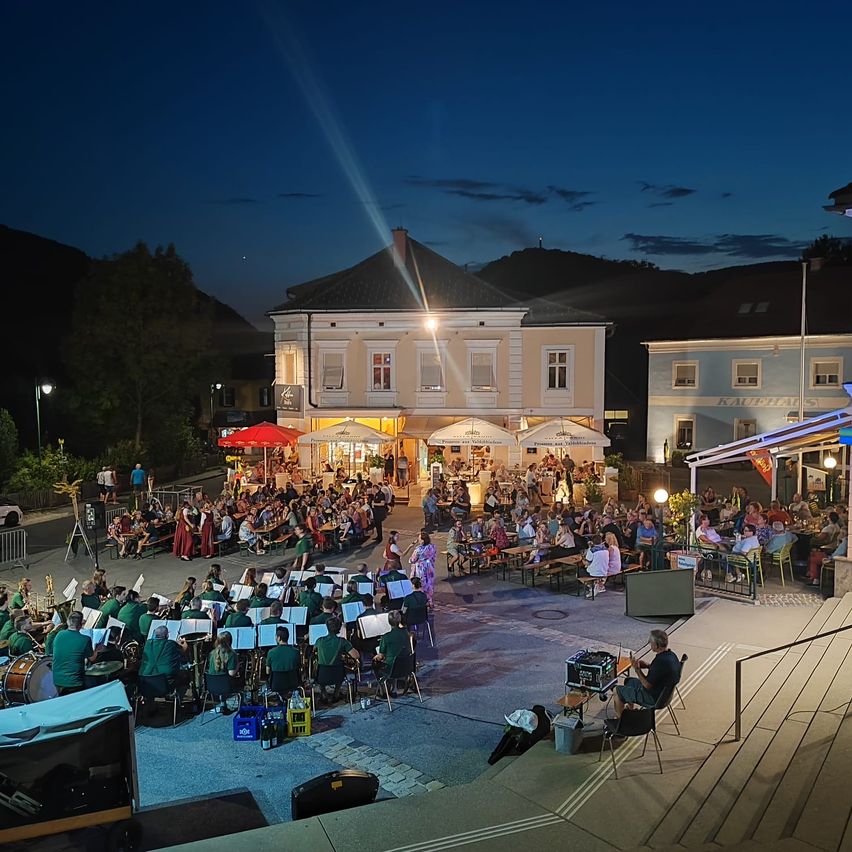 Ein Orchester spielt bei Nacht vor einer großen Menschenmenge im Freien, mit Gebäuden und Bergen im Hintergrund.