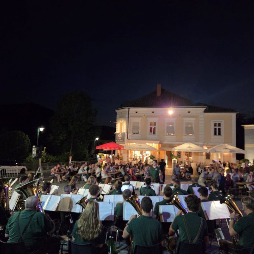 Ein Blasorchester spielt bei Nacht vor einem großen Gebäude, vor dem sich viele Menschen versammelt haben, um zuzuschauen. Die Bandmitglieder sind in grünen Uniformen gekleidet und spielen verschiedene Blechblasinstrumente. Das Gebäude hat mehrere Fenster und wird von Lichtern beleuchtet.