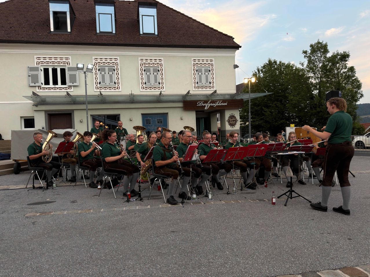 Ein Blasorchester spielt im Freien vor einem Gebäude mit dem Schild Dorfgasthof. Die Musiker sitzen und spielen verschiedene Instrumente.