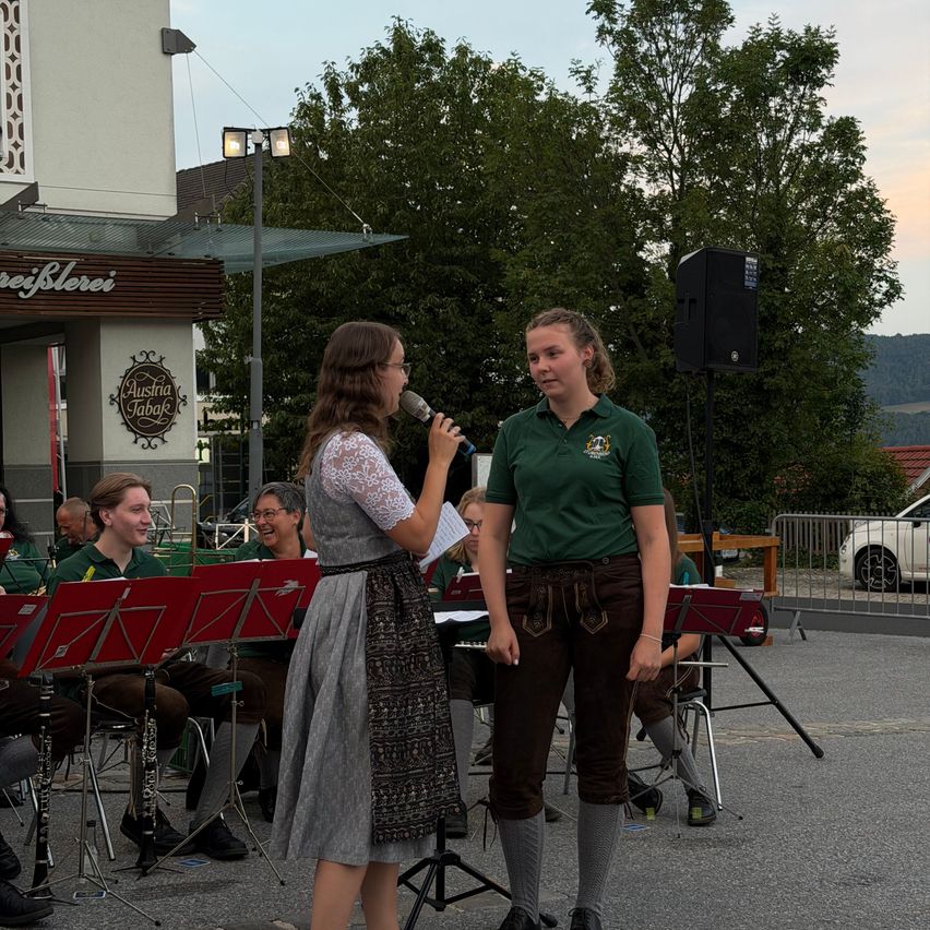 Zwei Frauen stehen vor einem Mikrofon, eine hält ein Musikblatt in der Hand, während eine Band hinter ihnen vor einem Gebäude spielt.