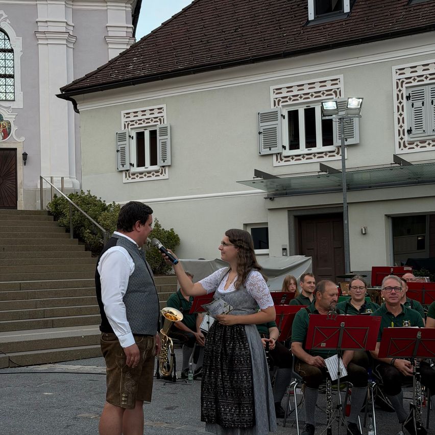 Ein Mann und eine Frau stehen vor einem Gebäude, hinter ihnen steht eine Brassband. Die Frau hält ein Mikrofon und spricht mit dem Mann.