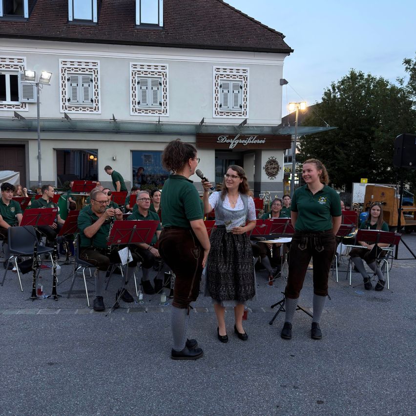 Eine Gruppe von Musikern in grüner Kleidung spielt auf einer Straße vor einem Gebäude mit dem Schild Dorfgeflohneri.