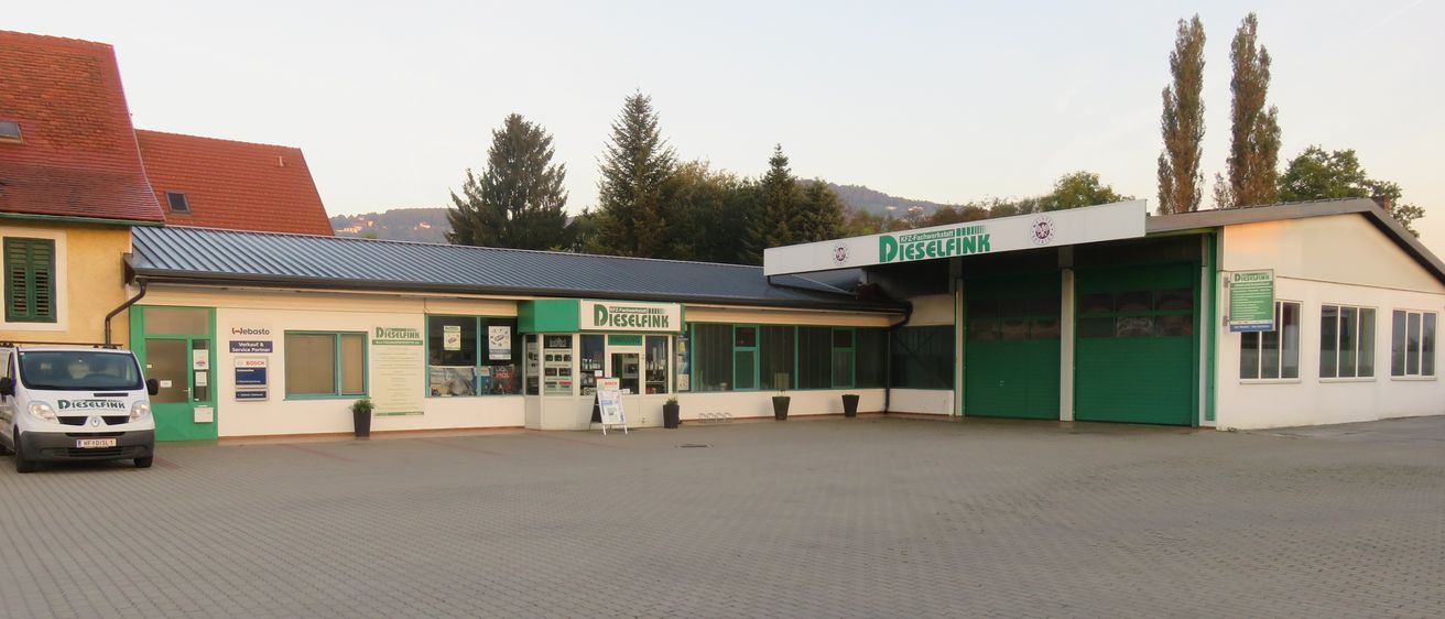 Ein Außenansicht eines Gebäudes mit der Aufschrift Dieselfink, mit einem grauen Dach, grünen Fenstern und mehreren Topfpflanzen. Bäume und Berge im Hintergrund.