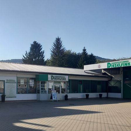 Außenansicht eines Dieselfink-Ladens mit einem Schild oben und Bäumen dahinter. Vorne stehen Topfpflanzen und ein Schild.