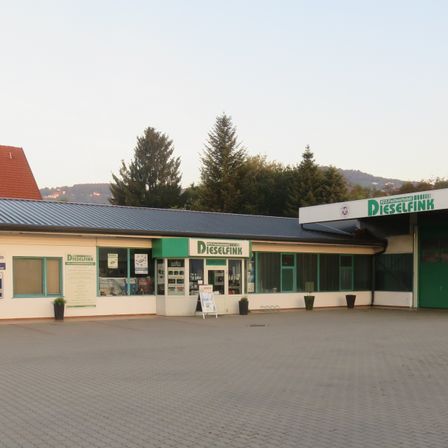 Ein Gebäude mit einem Metalldach und einem grünen Schild mit der Aufschrift 'Dieselflink', mit mehreren Fenstern und einem Schild davor. Bäume und ein Haus sind im Hintergrund zu sehen.