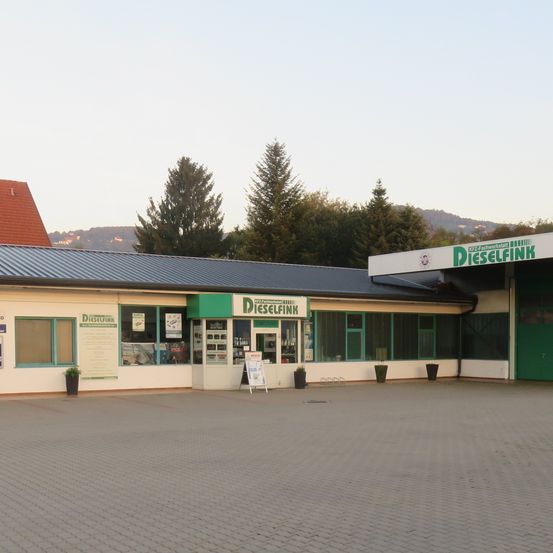 Ein Gebäude mit einem Metalldach und einem grünen Schild mit der Aufschrift 'Dieselflink', mit mehreren Fenstern und einem Schild davor. Bäume und ein Haus sind im Hintergrund zu sehen.
