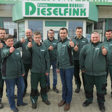 Eine Gruppe von Mitarbeitern in grünen Uniformen steht vor einem Geschäft namens Firma Dieselfink, lächelt und gibt Daumen hoch.