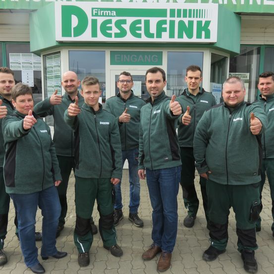 Eine Gruppe von Mitarbeitern in grünen Uniformen steht vor einem Geschäft namens Firma Dieselfink, lächelt und gibt Daumen hoch.