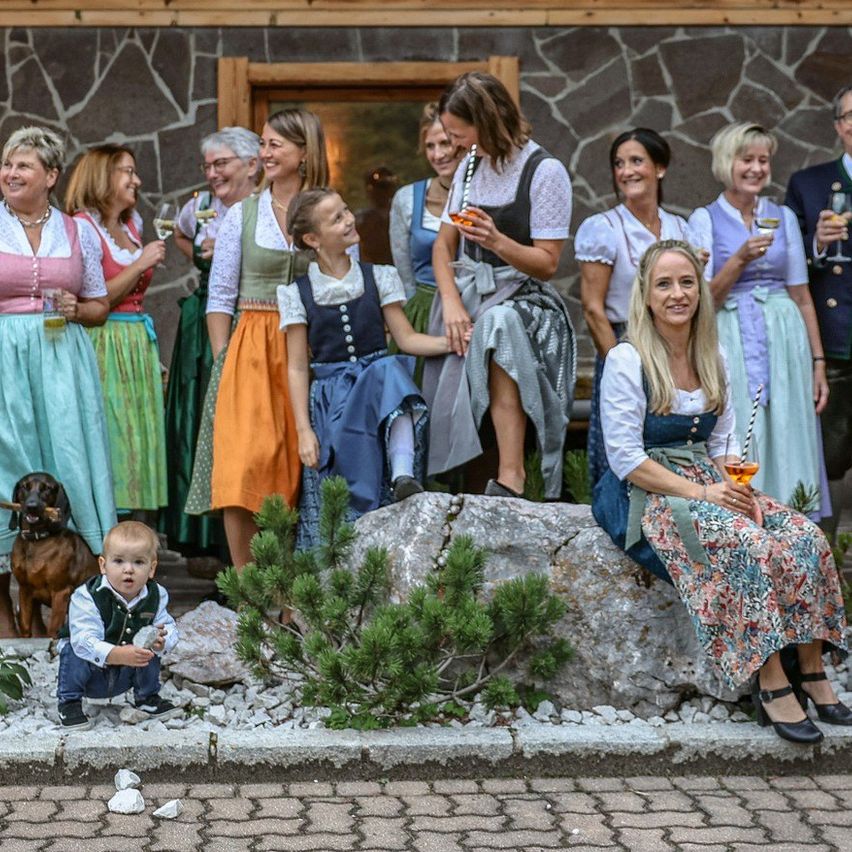 Eine Gruppe von Menschen in traditioneller Kleidung, einige sitzend und andere stehend, posiert für ein Foto vor einem Holzgebäude. Ein Kleinkind und ein Hund sind ebenfalls anwesend.
