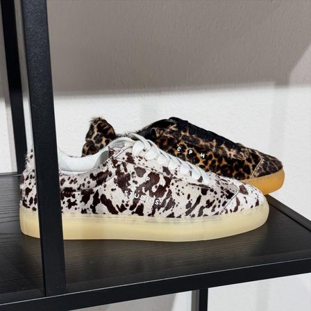 Ein Paar Leopardenmuster-Sneaker und ein Paar Kuh-Print-Sneaker werden auf einem Regal ausgestellt. Beide Schuhe haben weiße Sohlen.
