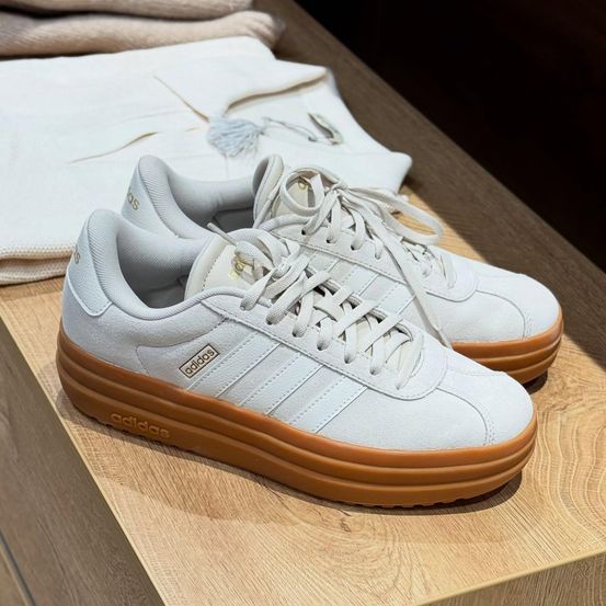 Ein Paar weiße Adidas-Sneaker mit braunen Sohlen auf einem Holztisch mit einem weißen Tuch.