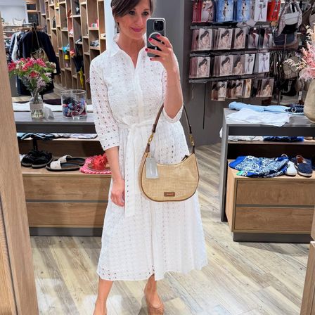 Eine Frau macht ein Selfie in einem Bekleidungsgeschäft, trägt ein weißes Kleid und eine braune Handtasche. Hinter ihr sind Regale mit verschiedenen Kleidungsstücken zu sehen, und an einem Tresen sind Schuhe und Accessoires ausgestellt.