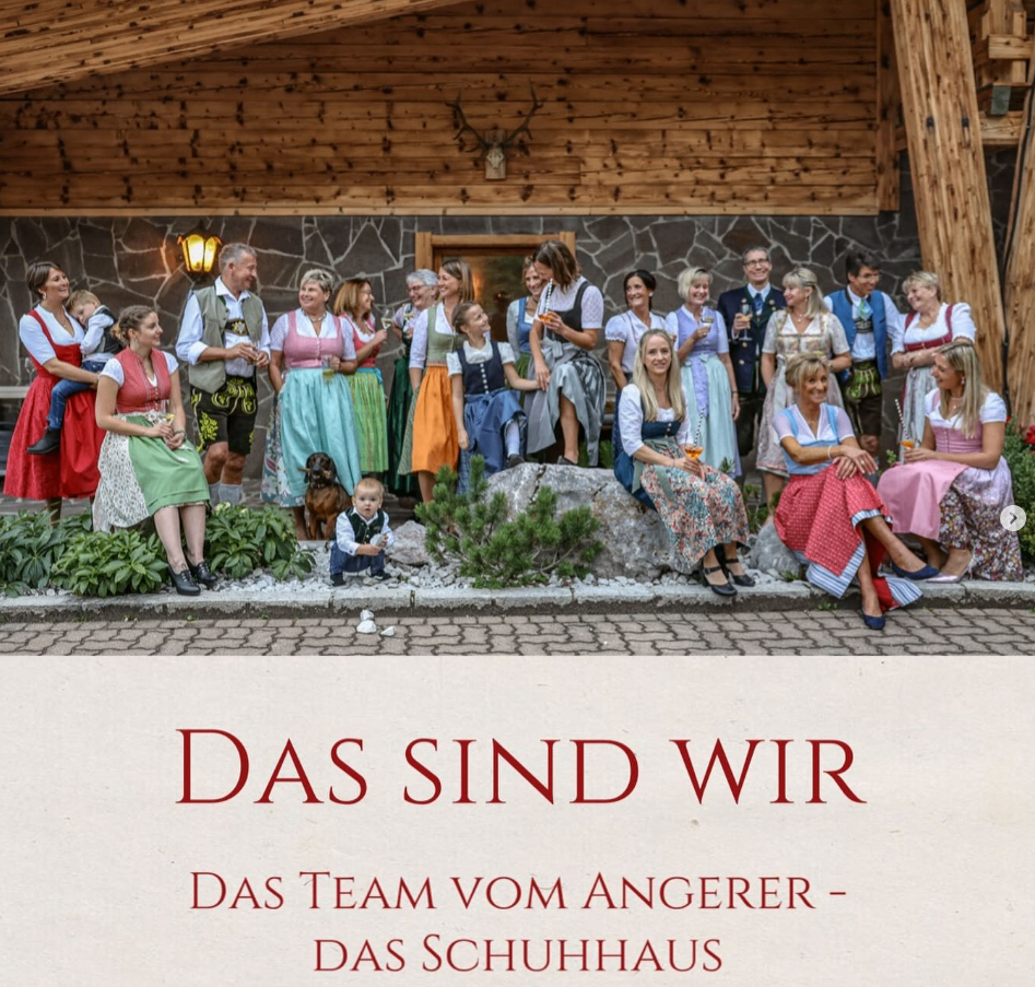 Group of adults and children in traditional Bavarian clothing standing in front of a building with stone walls. A baby is sitting on the ground. Text reads, 'DAS SIND WIR, DAS TEAM VOM ANGERER - DAS SCHUHHAUS'.