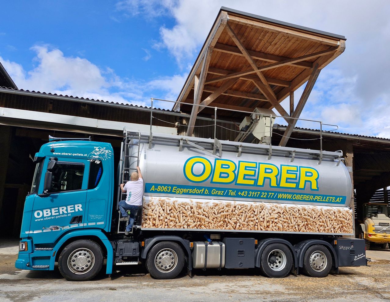 Ein blauer Lkw mit der Aufschrift 'Oberer' steht vor einer Holzkonstruktion. Ein Mann klettert auf der Leiter des Lkw. Die Seite des Lkw ist mit einem Haufen brauner Holzpellets gefüllt.